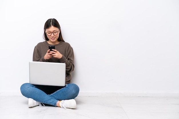 Молодая женщина с ноутбуком сидит на полу, отправляя сообщение с мобильного телефона