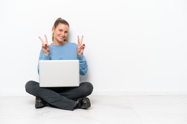 Giovane donna con un computer portatile che si siede sul pavimento che mostra il segno di vittoria con entrambe le mani