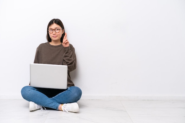 Young woman with a laptop sitting on the floor with fingers crossing and wishing the best