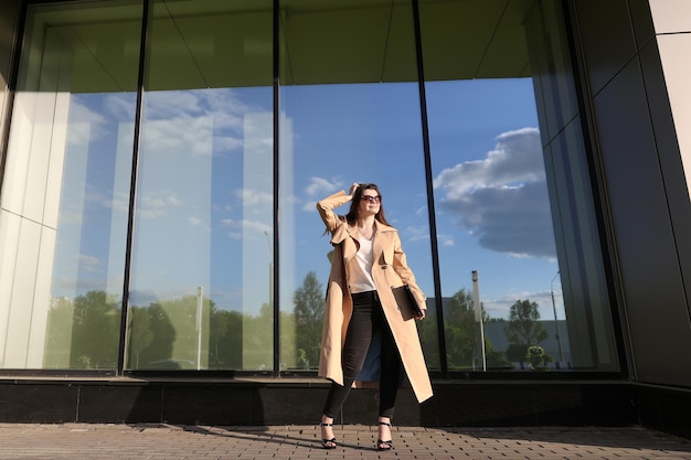 A young woman with a laptop walking through city streetBusiness woman in stylish trench coat outside