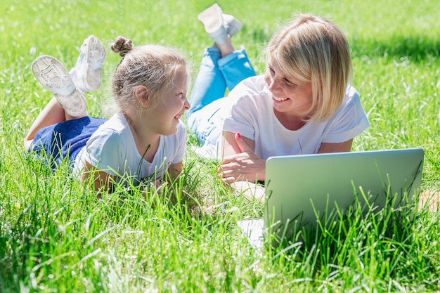 Young woman with a little daughter lie in the park with a laptop and laugh. Blogging, online training and distance learning.