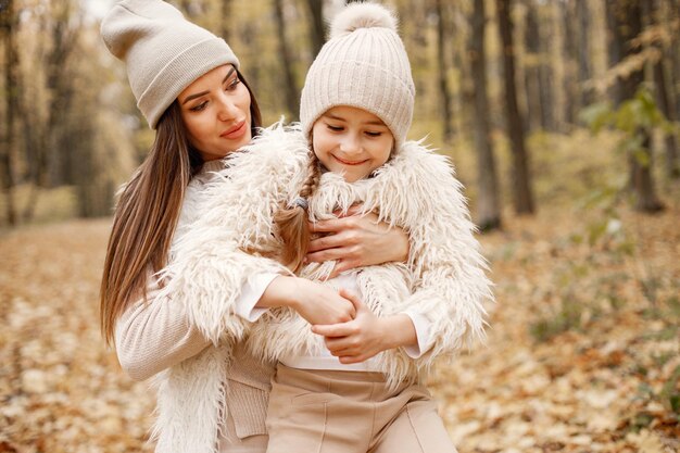 Young woman with little girl standing in autumn forest. Brunette woman play with her daughter. Girl wearing beige sweater and mother wearing white clothes.