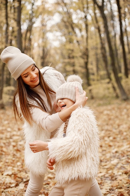 Young woman with little girl walking in autumn forest. Brunette woman play with her daughter. Girl wearing beige sweater and mother wearing white clothes.