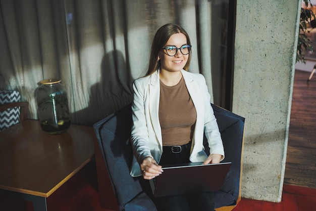 A young woman with long blond hair in stylish clothes is sitting in a chair and working on a laptop Work in a modern office with large windows