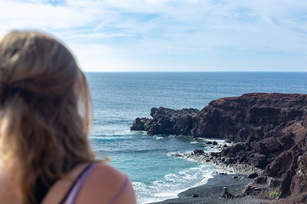 Foto giovane donna con lunghi capelli biondi seduti sulla spiaggia e guardando l'orizzonte. oceano blu e costa rocciosa.