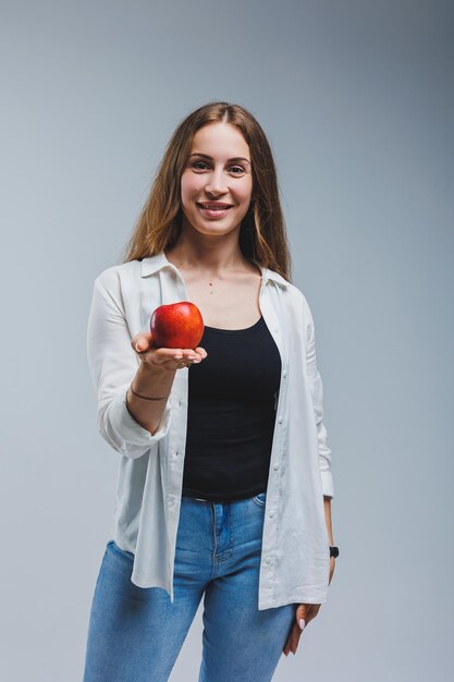 A young woman with long brunette hair dressed in a white shirt and jeans holds a fresh red apple in her hands Healthy food concept White background