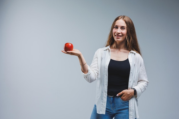 A young woman with long brunette hair dressed in a white shirt and jeans holds a fresh red apple in her hands Healthy food concept White background