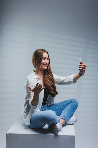 Young woman with long brunette hair wearing a white shirt and jeans having a video call conversation on the phone A woman in jeans and a plain white shirt with a mobile phone White background