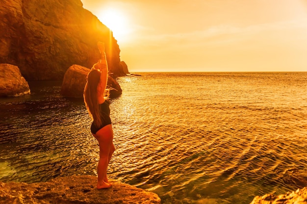 Молодая женщина с длинными волосами в черном купальнике и браслетах в стиле бохо практикует на открытом воздухе на коврике для йоги у моря на закате. Женская йога-фитнес. Гармония здорового образа жизни и медитация.