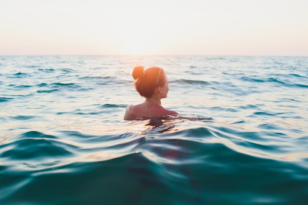 Giovane donna con i capelli lunghi, bionda, in topless, seduto in acqua e con in mano un bikini sotto il sole.