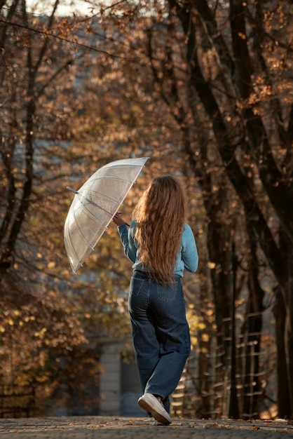 Young woman with long hair in blue sweater and pants walking\
with transparent umbrella in autumn park back view