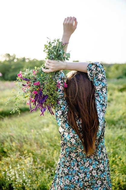 A young woman with long hair in a colorful short dress holds a bouquet of wild flowers in her raised...