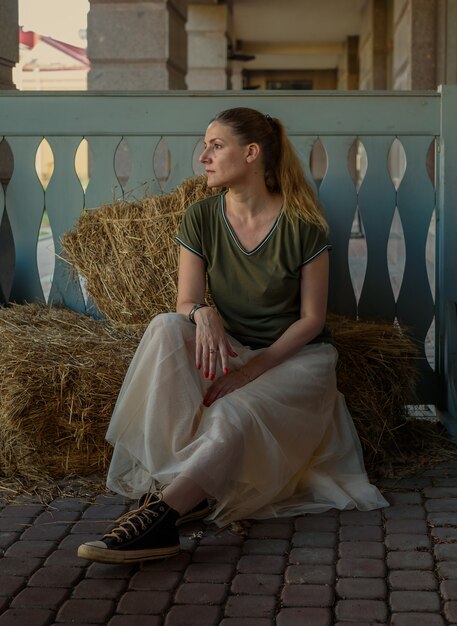 Молодая женщина с длинными волосами, в пышной юбке, футболке сидит летом на соломе в экологическом кафе. Портрет женщины в естественной сельской местности. Концепция экологического экотуризма.