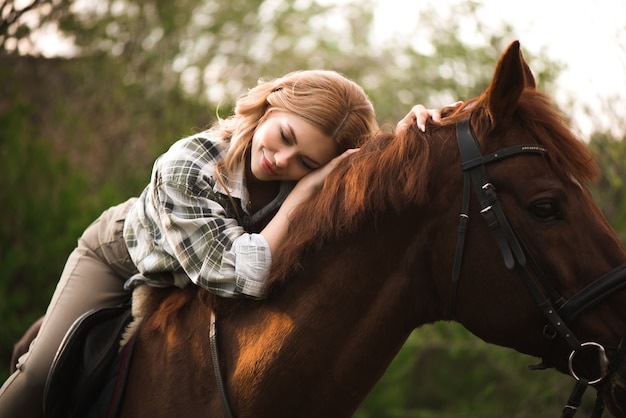 Young woman with long hair posing with a brown horse in a forest in a sunny meadow.