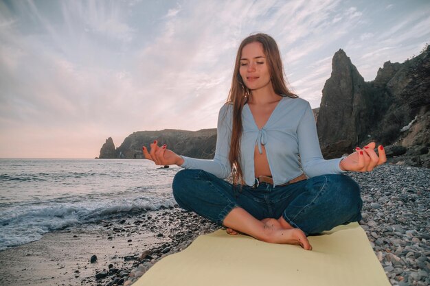 Young woman with long hair practicing stretching outdoors on yoga mat by the sea on a warm sunset