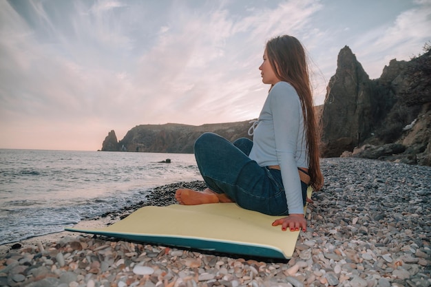 暖かい夕日の海沿いのヨガマットで屋外ストレッチを練習している長い髪の若い女性