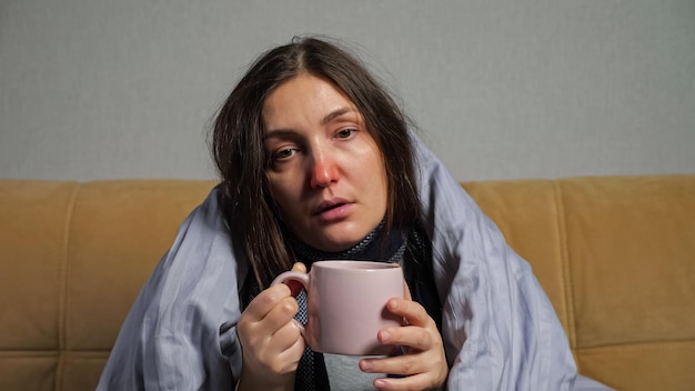 Young woman with long hair and runny nose falls ill and drinks\
hot tea from mug wrapped in warm blanket, sitting on comfortable\
sofa in living room.