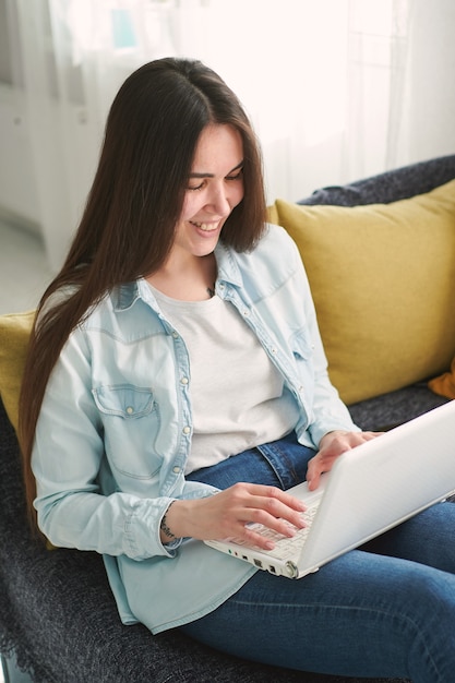 Young woman with long hair sits on the couch at home and works on laptop