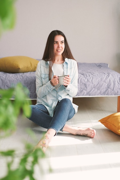 Young woman with long hair sits on the floor at home and drinks coffee