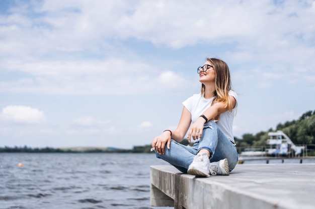 Young woman with long hair in stylish glasses posing on the concrete shore near the lake. Girl dressed in jeans and t-shirt smiling and looking away