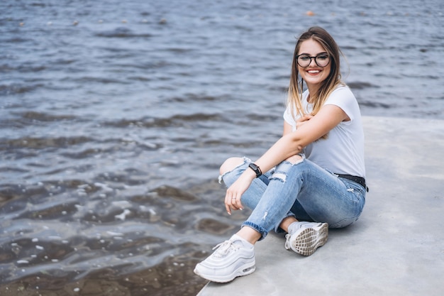 Молодая женщина с длинными волосами в стильных очках позирует на бетонном берегу у озера