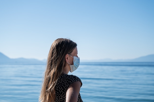 Молодая женщина с длинными волосами в защитной медицинской маске стоит у моря, глядя вдаль