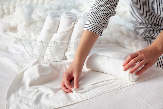 Young woman with long red hair is sitting on bed and folding clean white hand towels into mesh basket in bedroom. Stack of roll up towels with copy space. Neat and tidy linen. Hypoallergenic textile.