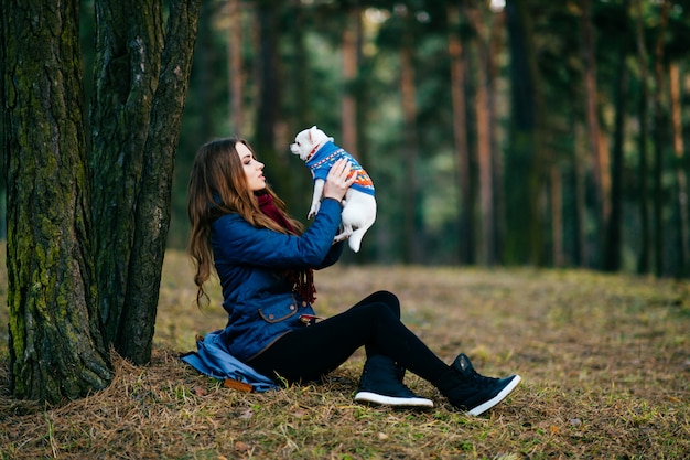 Young woman with long sitting on ground beyond trees in forest with her lovely chihuahua puppy in hands.