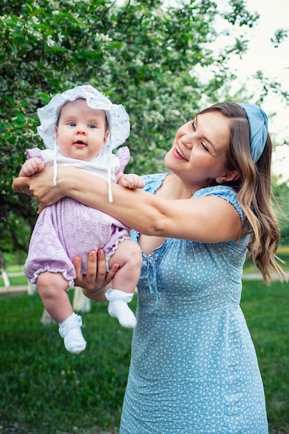 Young woman with loose fair hair rocks in arms baby girl and\
plays with kid standing in park against green trees in white\
blossom