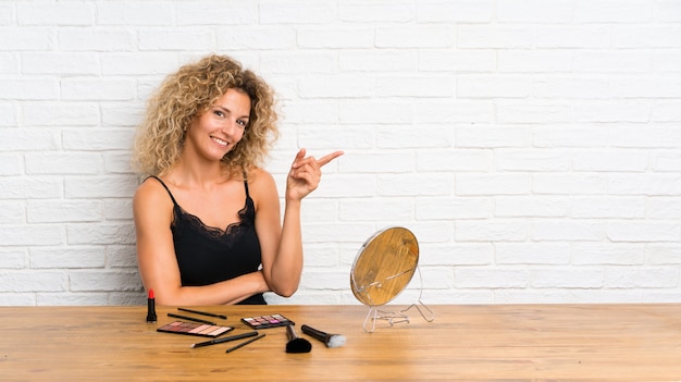 Young woman with lots of makeup brush in a table pointing finger to the side