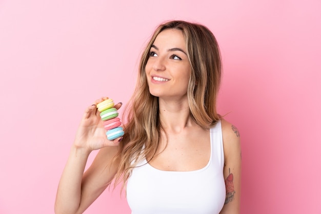 Young woman with  macarons over isolated pink wall looking up while smiling