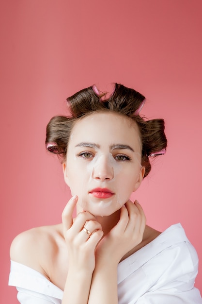 Young woman with a mask and curlers touching her face.