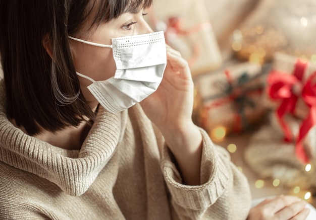 Young woman with a mask on her face in a cozy beige sweater. The concept of christmas during the coronavirus.