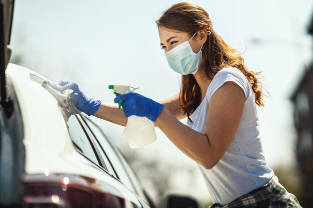 A young woman with a mask on her face and protective gloves on her hands, wipes her car holding a cloth in one and a bottle with disinfectant in the other hand.