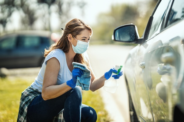 A young woman with a mask on her face and protective gloves on her hands, wipes her car holding a cloth in one and a bottle with disinfectant in the other hand.