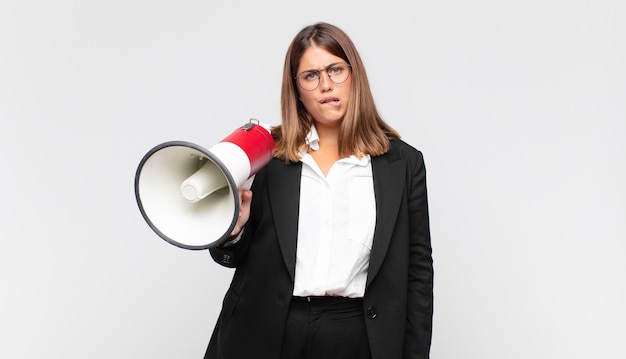 Young woman with a megaphone looking puzzled and confused, biting lip with a nervous gesture, not knowing the answer to the problem