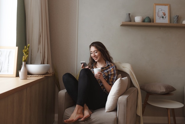 Una giovane donna con un telefono cellulare è seduta su una comoda sedia vicino alla finestra di casa e sta bevendo il tè. interior design per la casa. libero professionista