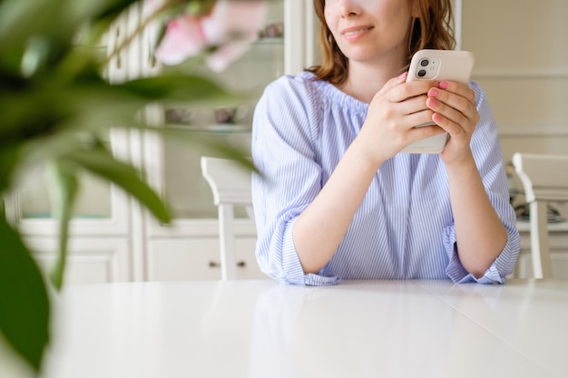 Young woman with modern mobile phone sits at white table smiling behind blurred flowers on foregroun