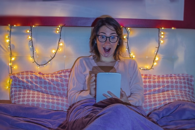 Photo young woman with mouth open using digital tablet while sitting on bed at home