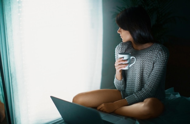 Photo young woman with mug sitting by laptop on bed at home