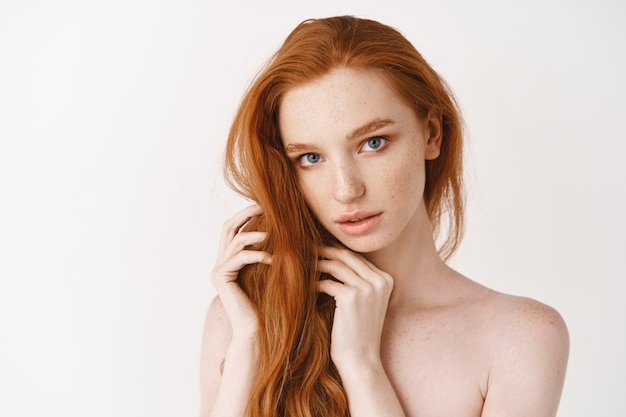 Фото Молодая женщина с натуральными блестящими рыжими волосами и идеальной бледной кожей, смотрящая вперед, чувственным взглядом, стоит обнаженной над белой стеной
