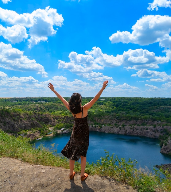 A young woman with open hands up stands on a hill above a picturesque lake. Freedom concept. Stock phototography.