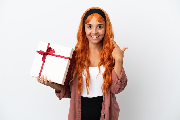 Молодая женщина с оранжевыми волосами держит подарок на белом фоне, показывая жест рукой вверх