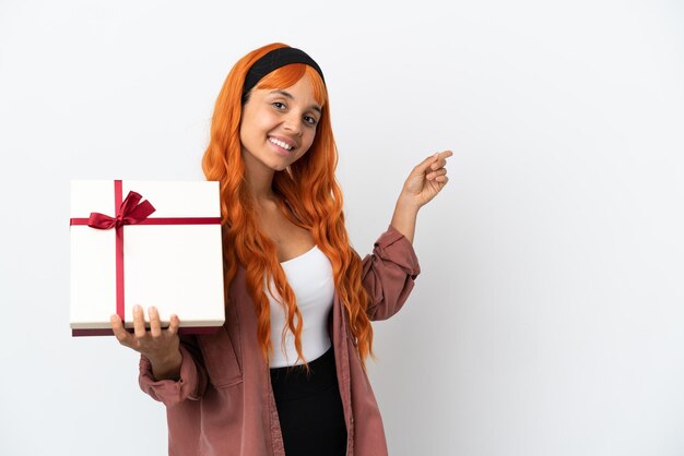 Foto giovane donna con i capelli arancioni che tiene un regalo isolato su sfondo bianco che punta indietro