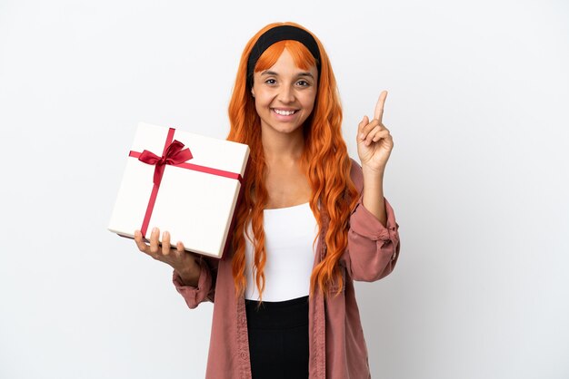 Молодая женщина с оранжевыми волосами держит подарок на белом фоне, показывая и поднимая палец в знак лучших