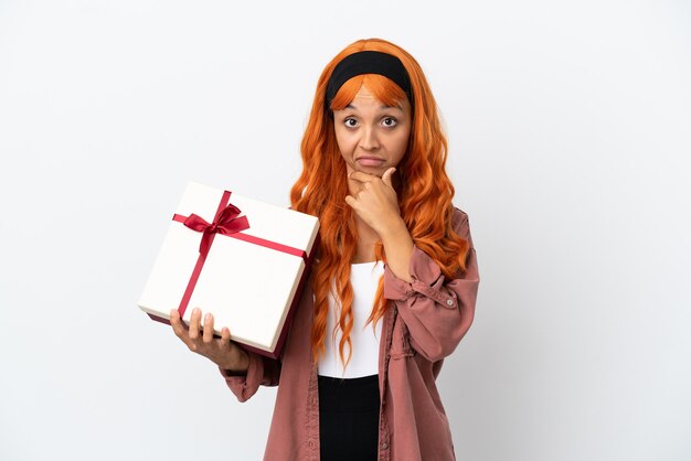 Giovane donna con i capelli arancioni che tiene un regalo isolato su sfondo bianco pensando