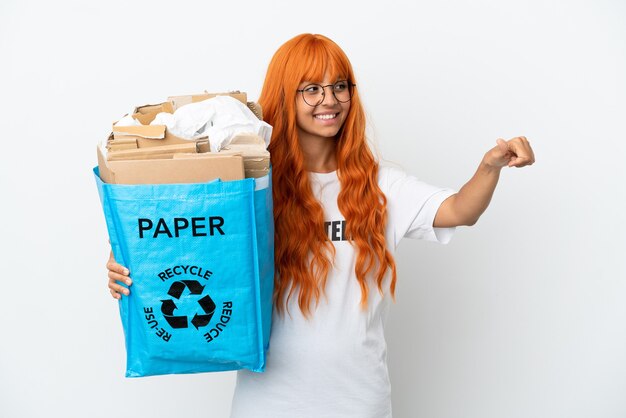 Young woman with orange hair holding a recycling bag full of paper to recycle isolated on white background giving a thumbs up gesture