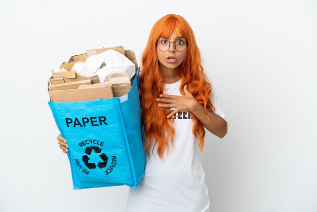 Young woman with orange hair holding a recycling bag full of paper to recycle isolated on white background surprised and shocked while looking right