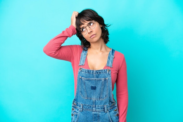 Young woman with overalls isolated background having doubts while scratching head