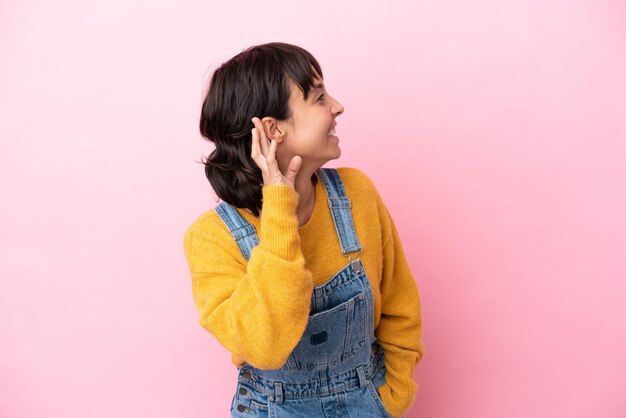 Young woman with overalls isolated background listening to something by putting hand on the ear